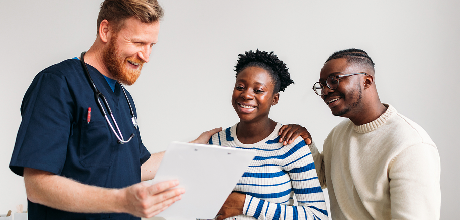 A nurse or doctor sharing a medical chart and smiling with a couple. Everyone looks to be happy and smiling while they read the chart.