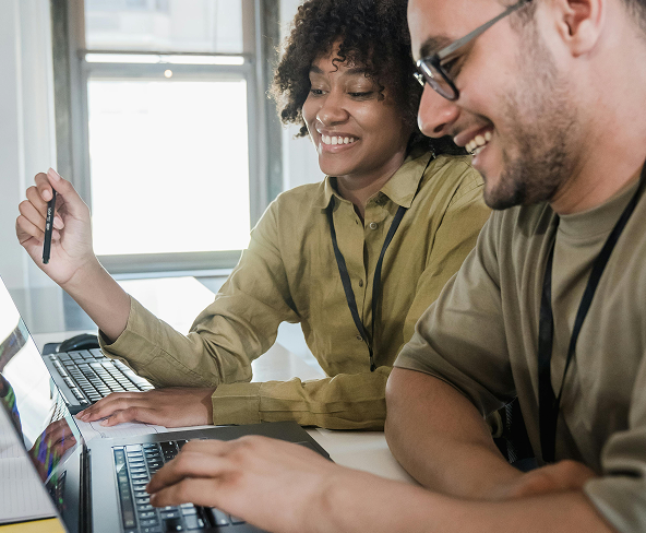 Two co-workers sitting at a desk smiling as they look at a computer screen.