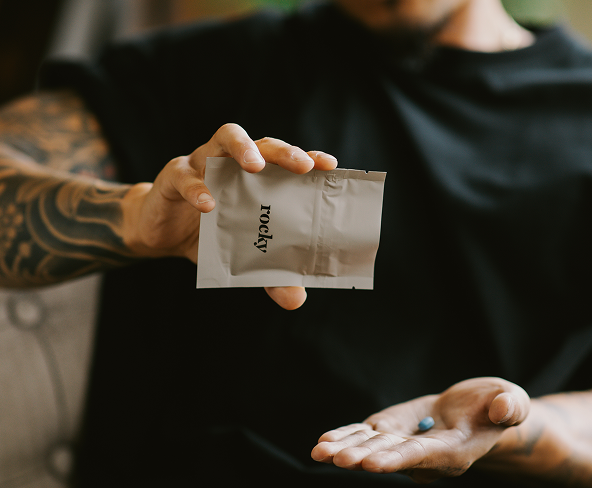 A close up view of a person holding a beige rocky package. A blue pill is in their hand.