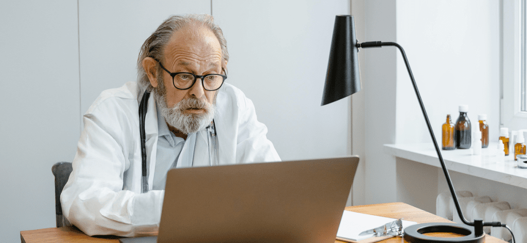 A very old doctor sitting at a desk and looking at his computer.