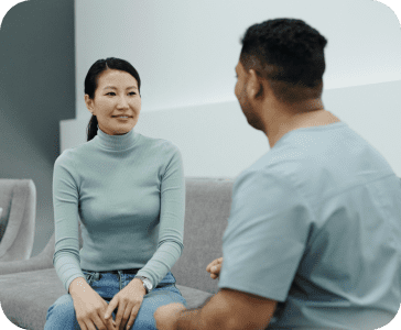 A woman and a man talking. The woman is smiling and facing towards us. They're both sitting on a grey couch.