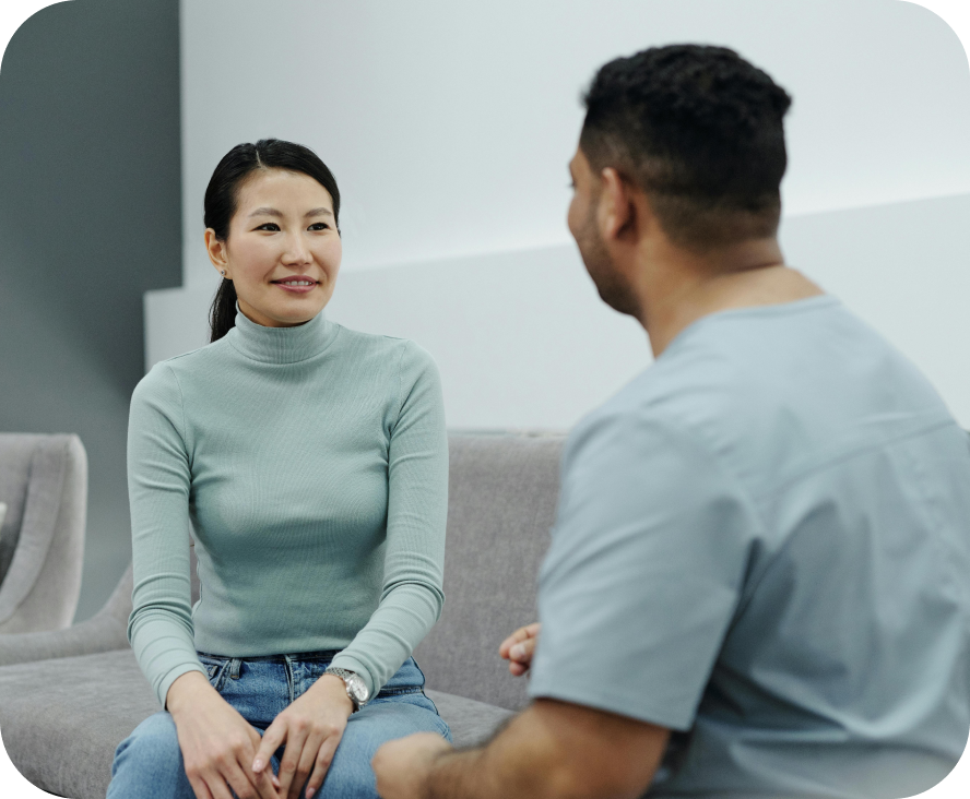 A woman and a man talking. The woman is smiling and facing towards us. They're both sitting on a grey couch.