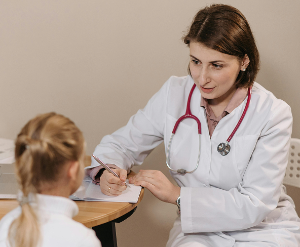 A doctor sitting down and talking to a child. The doctor is wearing a white coat with a red stethoscope