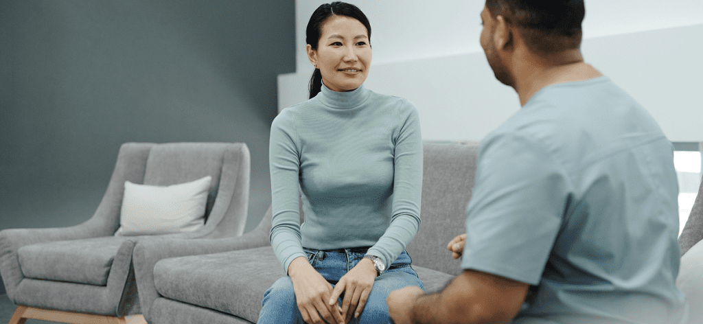 A woman and a man talking. The woman is smiling and facing towards us. They're both sitting on a grey couch.