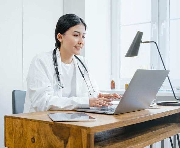 A doctor sitting at a wooden table reviewing notes on her computer.