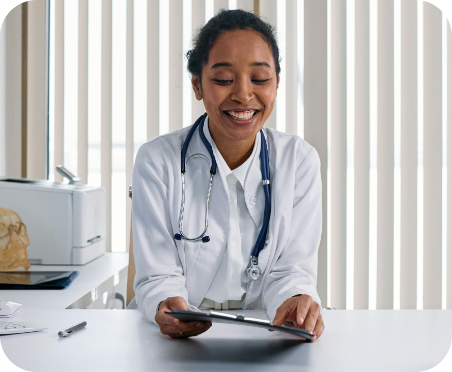 A doctor smiling while reading a medical chart. She's sitting down with a white lab coat on and a blue stethiscope