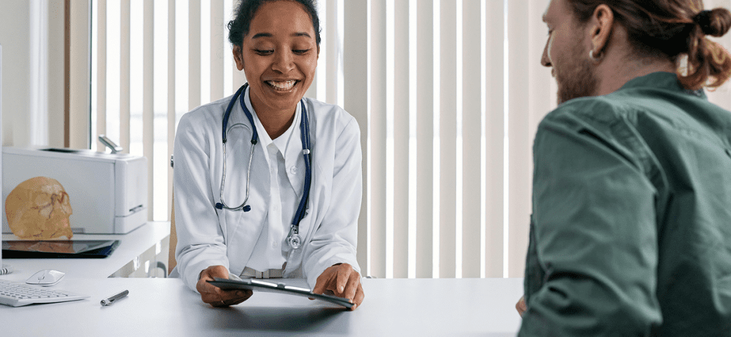 A doctor in her office sitting opposite a man in a green shirt. Both people are smiling as the doctor explains something on a chart.
