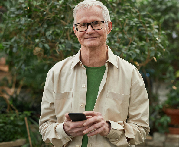 An older man smiling at the camera while he holds his phone.
